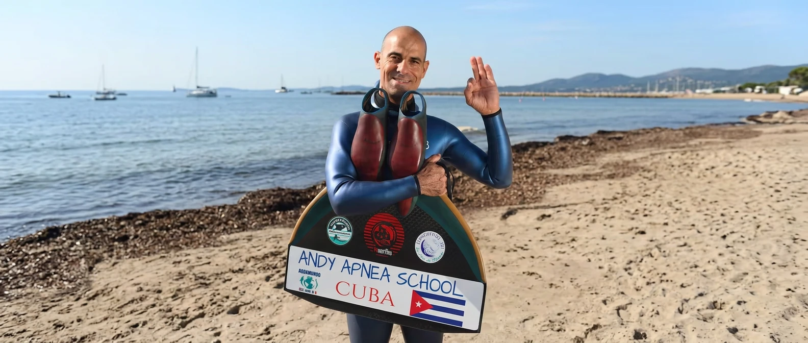 Andy Cabrera Ávila sostiene su monoaleta con el texto "Andy Apnea School Cuba" y la bandera cubana, en una playa con veleros al fondo.