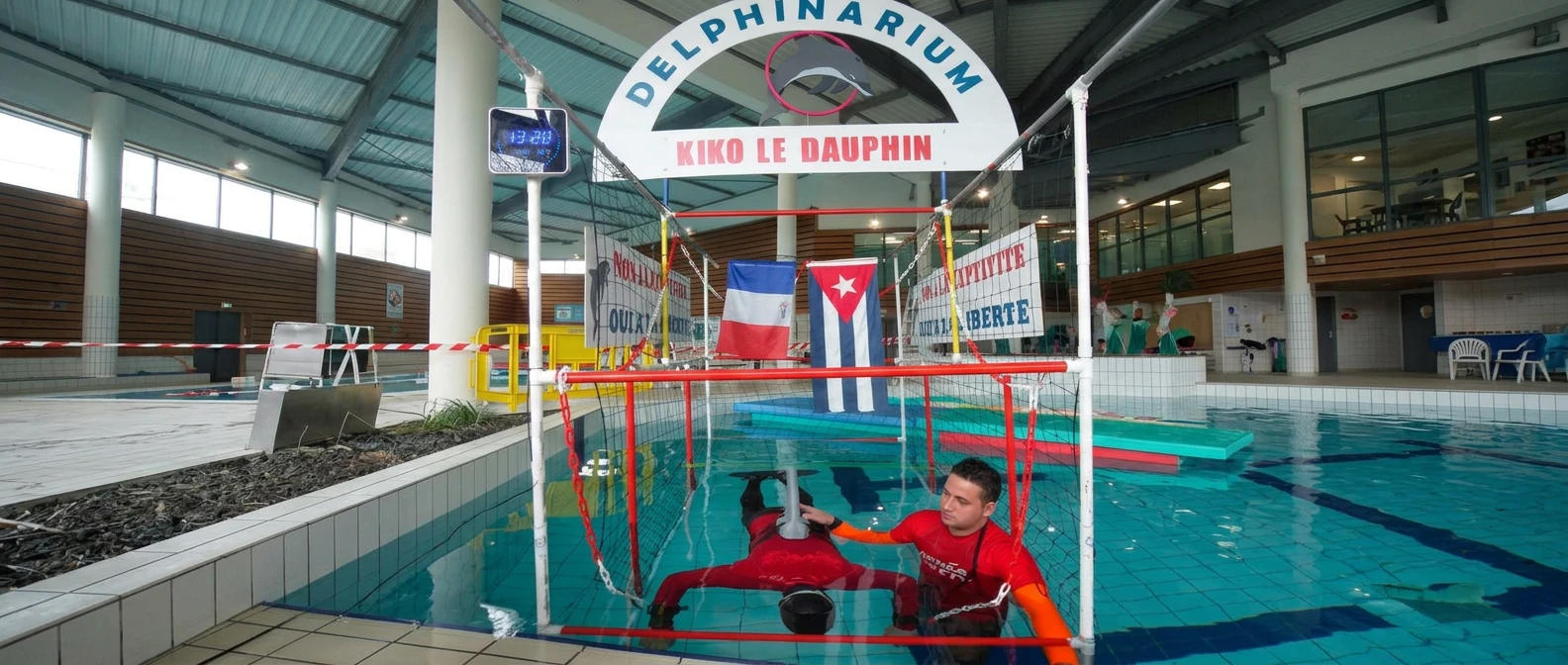 Un hombre dentro de una jaula sumergida en una piscina, bajo un cartel de "Delphinarium" y banderas de Francia y Cuba, con pancartas de "No a la cautividad, Sí a la libertad".