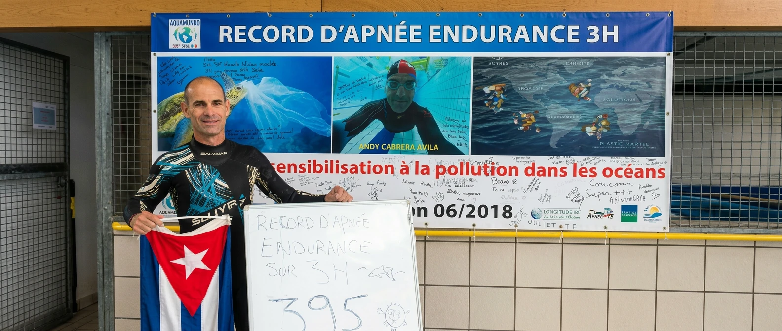 Andy Cabrera Ávila sostiene la bandera cubana junto a un cartel de "Récord d'apnée endurance 3H" sobre sensibilización contra la contaminación oceánica, con una pizarra que marca 395 largos.