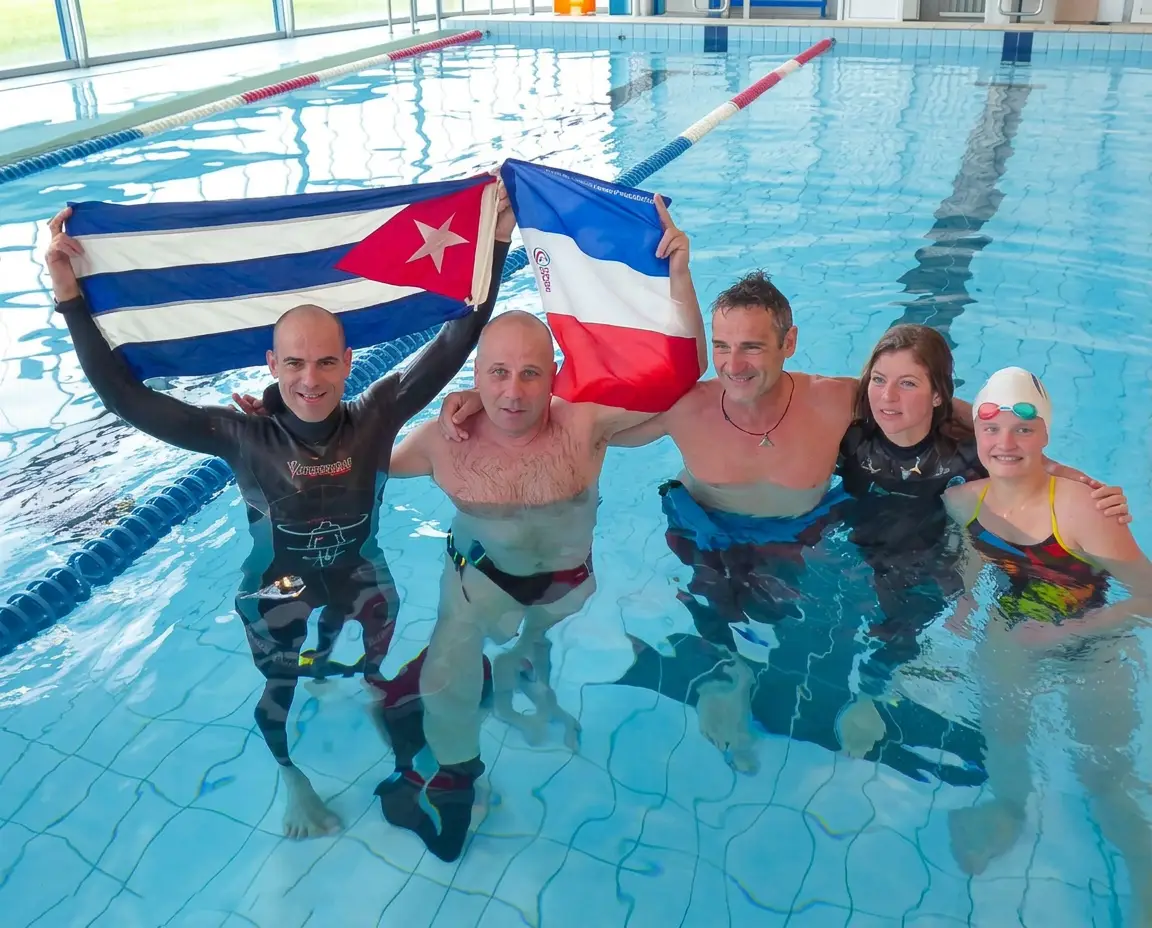 Cinq apnéistes en combinaison néoprène dans une piscine couverte brandissent les drapeaux de Cuba et de la France en signe de célébration.