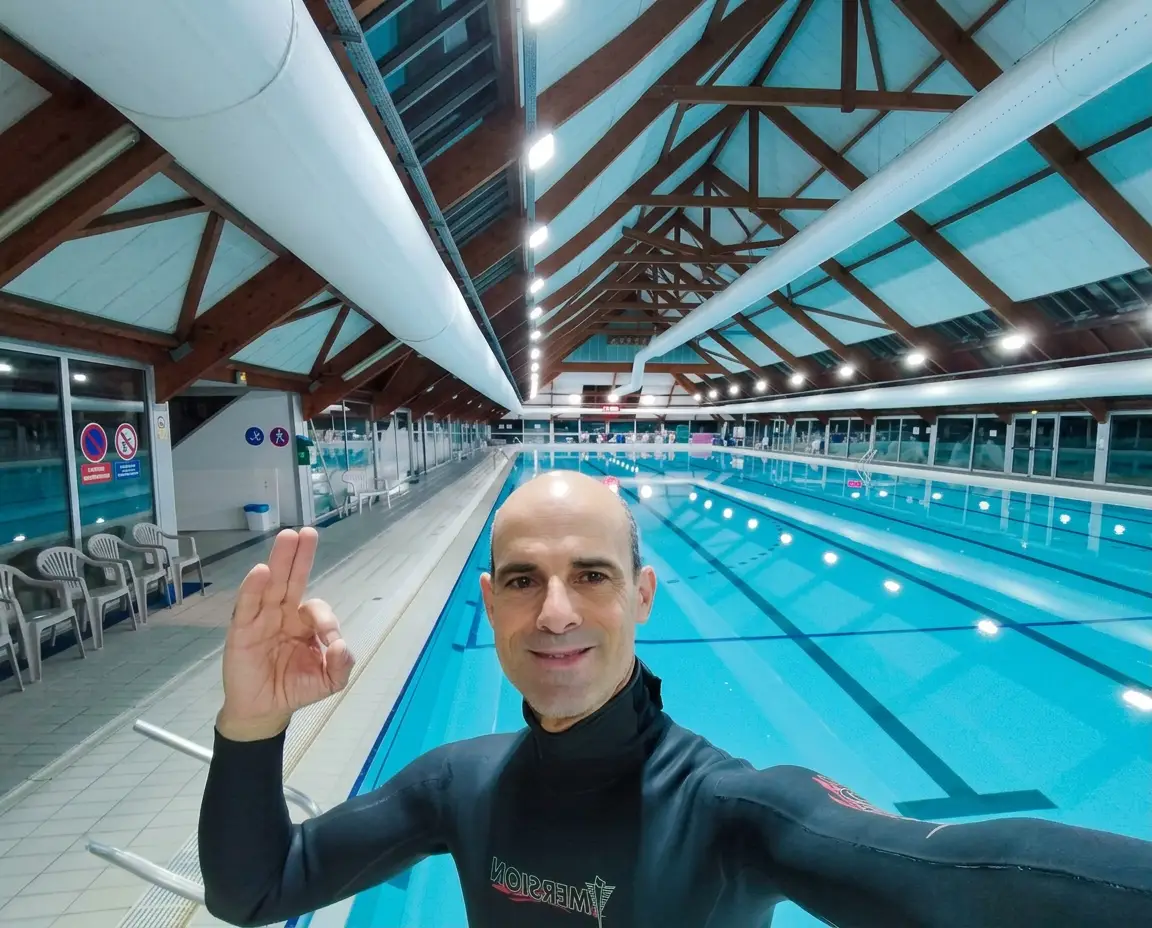 Un homme en combinaison néoprène fait un signe "ok" sur un selfie dans une piscine couverte avec des couloirs de nage.