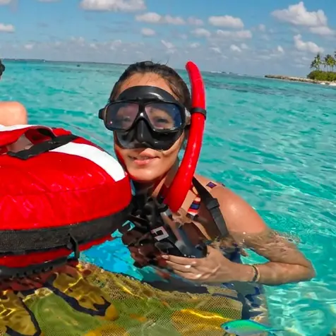 Mujer con máscara y tubo de snorkel rojo sonríe junto a una boya de señalización en aguas turquesas, con una isla al fondo.