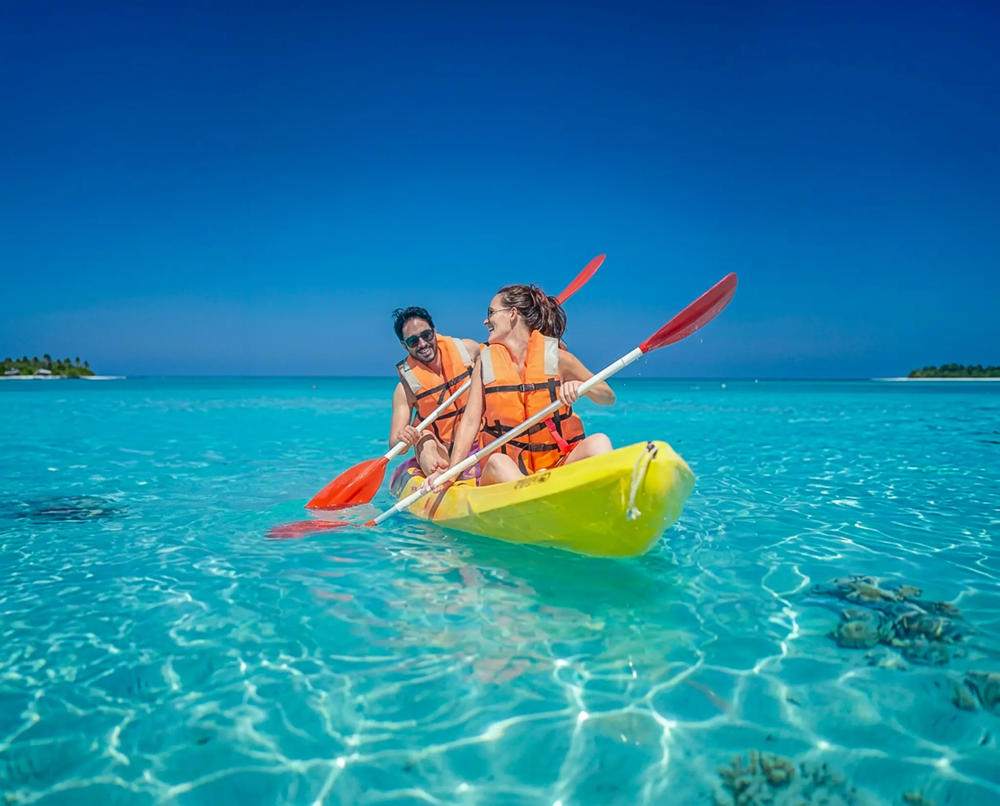 Pareja con chalecos salvavidas naranjas rema en un kayak amarillo sobre aguas turquesas cristalinas, con islas al fondo.