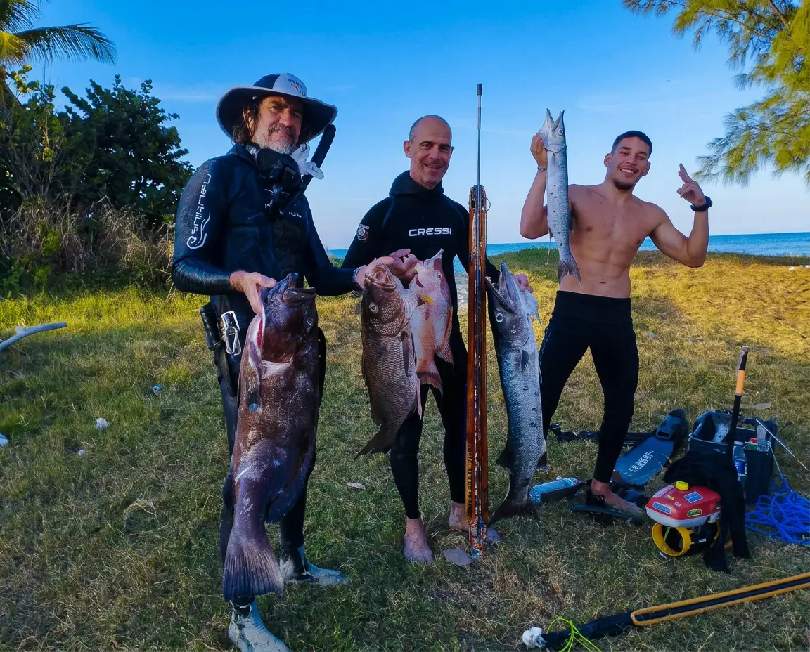 Trois apnéistes sur la plage montrent leur pêche sous-marine — mérou, vivaneau et barracuda — à côté de leur fusil et de leurs palmes.