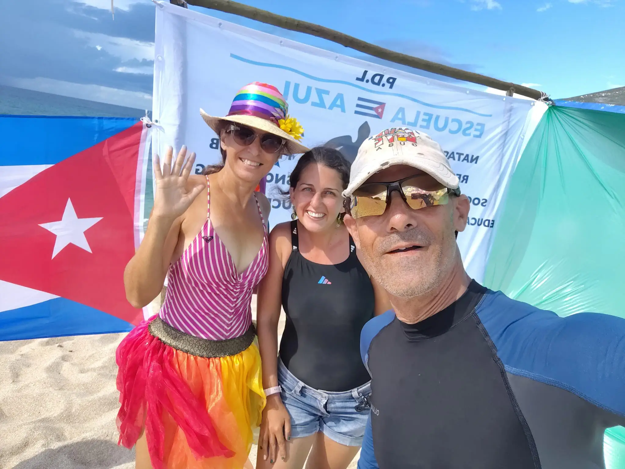 Tres integrantes de Escuela Azul se toman una selfie en la playa frente a la bandera de Cuba y el banner del proyecto.